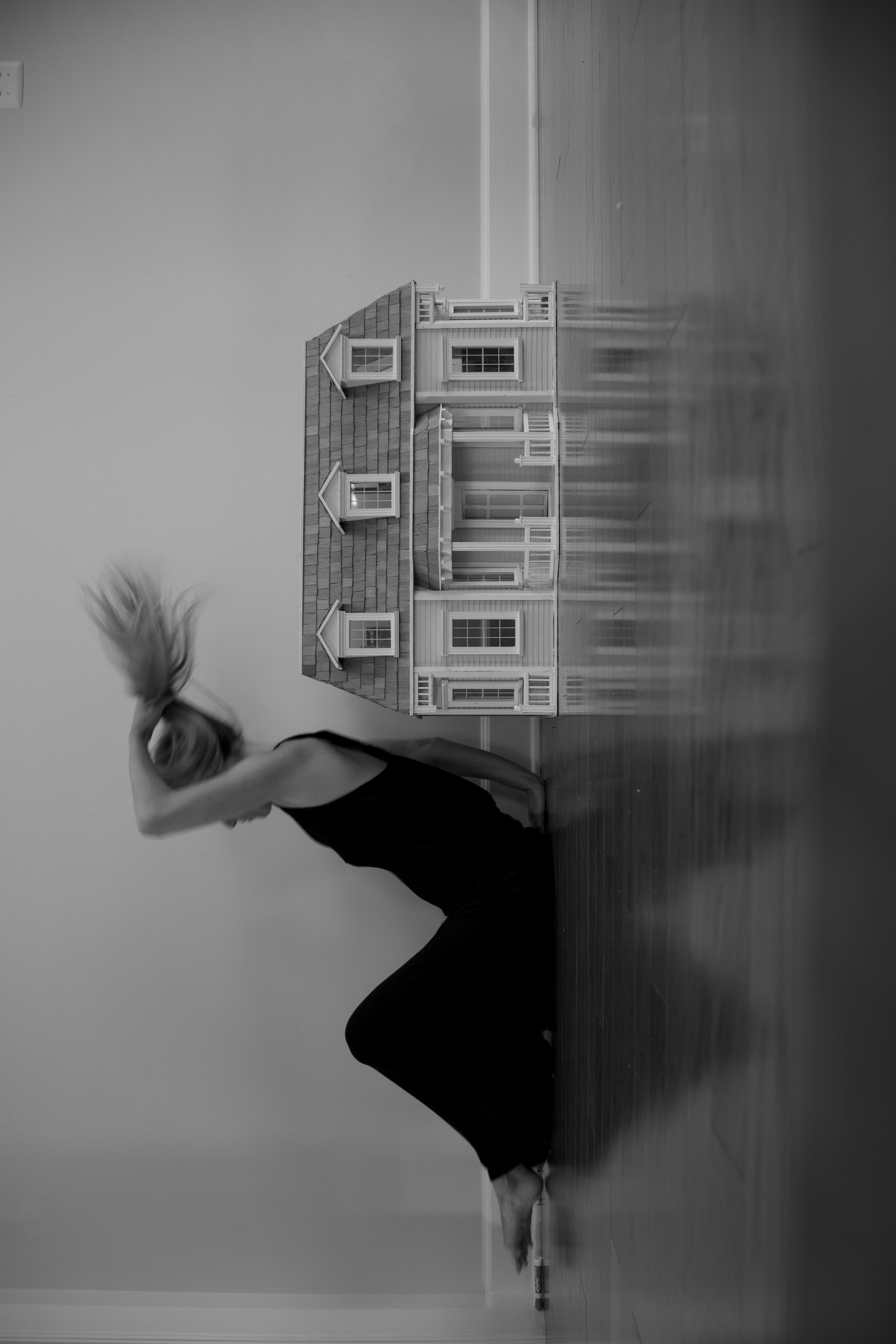 A woman bending beside a dollhouse in a blurred black-and-white interior